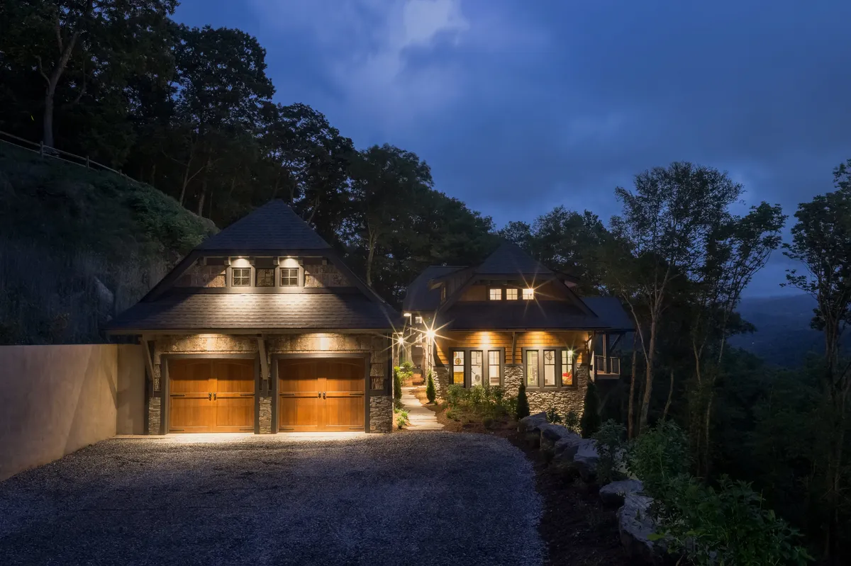 The warmly lit exterior of the house with illuminated garage doors and a landscaped pathway sets an inviting tone for cozy winter evenings. The image suggests stargazing, gathering around an outdoor fire, and peaceful moments of reflection, supporting the wintertime narrative.