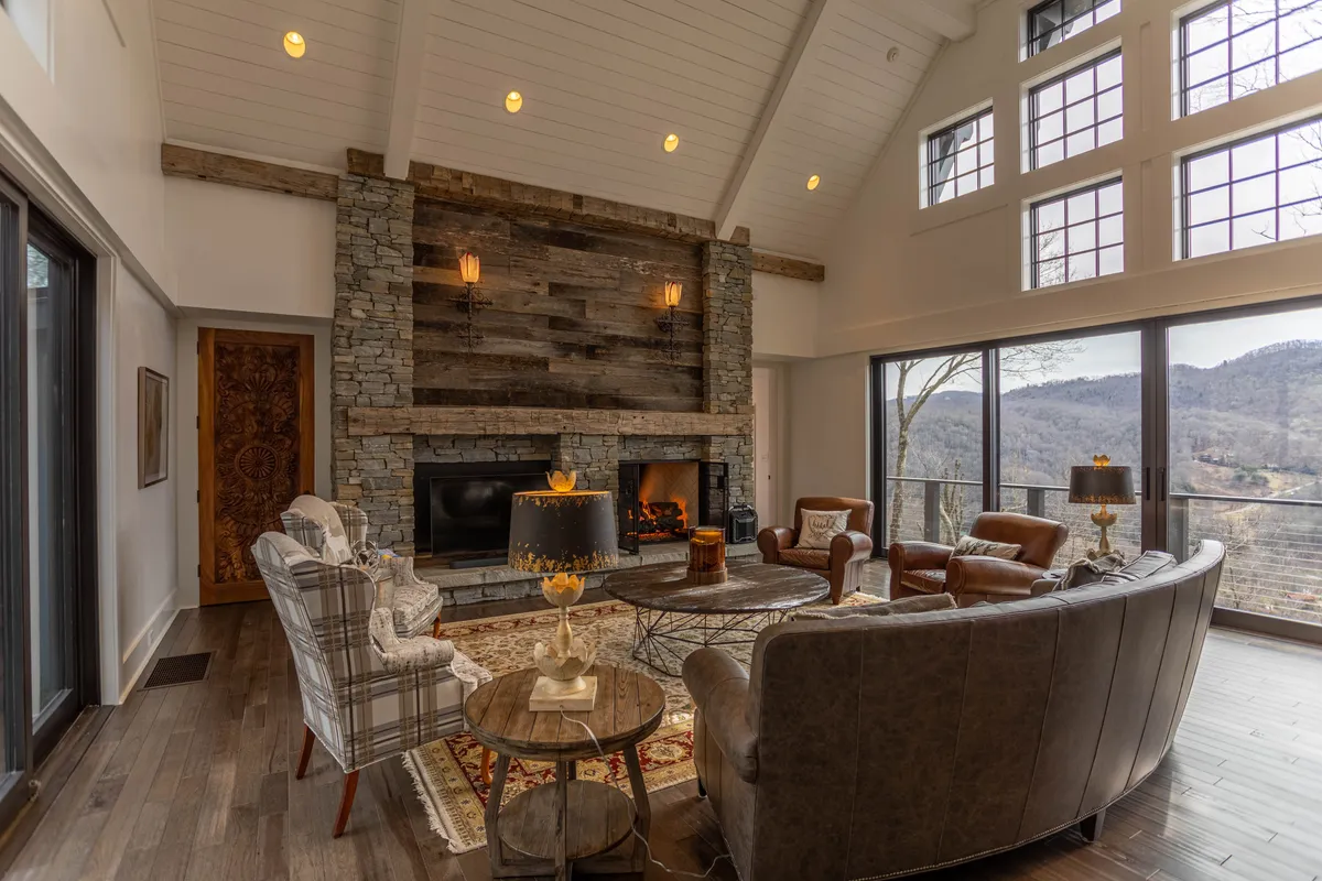 A spacious great room featuring a massive stone fireplace as the centerpiece, surrounded by plush, comfortable seating. Floor-to-ceiling windows frame striking mountain views, filling the space with natural light. The room radiates warmth, inviting guests to relax by the fire or enjoy the view, with hints of decor suggesting cozy gatherings and festive celebrations.