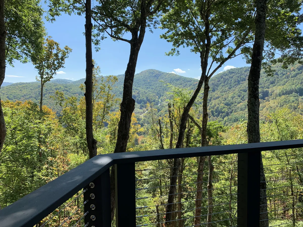 Captured from the deck, this image features breathtaking mountain vistas, a dense forest, and a clear sky. The deck railing in the foreground connects viewers to the immersive outdoor experience, emphasizing the privacy and natural beauty of the setting.