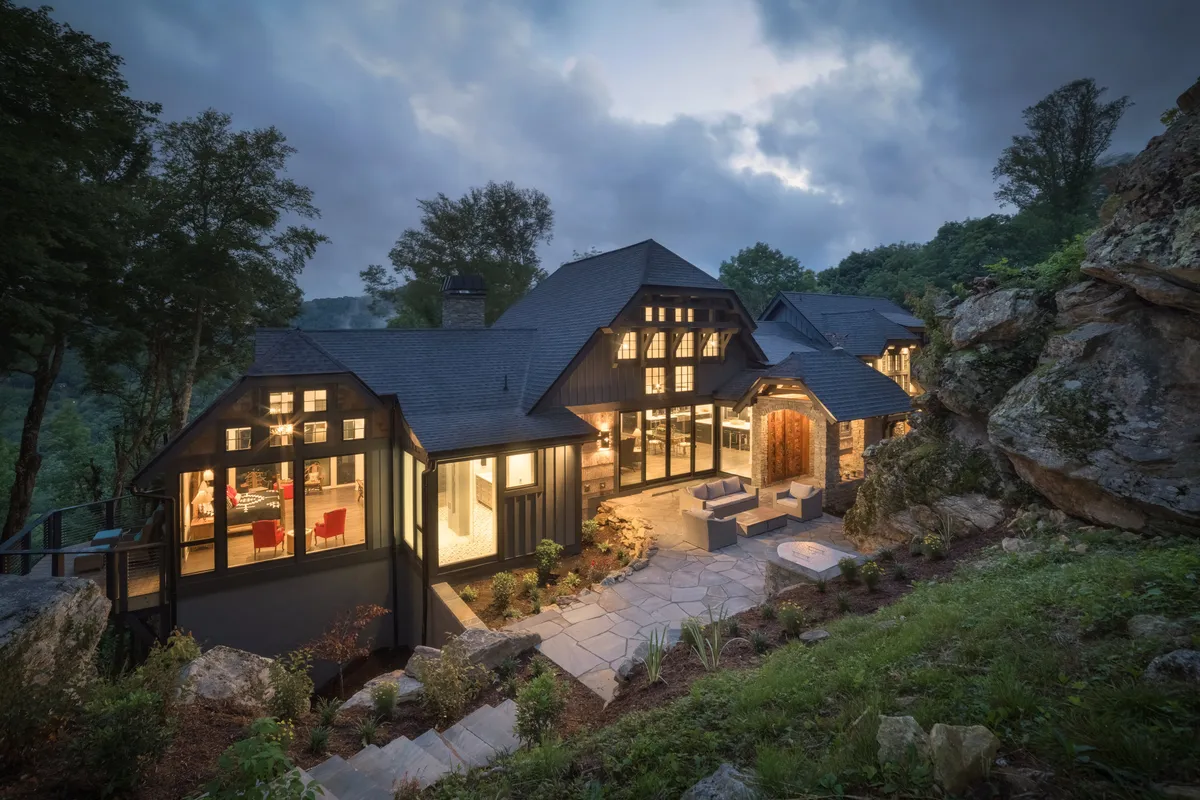 This exterior view showcases the stone pathway leading to the house, illuminated large windows hinting at the welcoming interior, an outdoor seating area for group gatherings, and the tranquil surrounding landscape. The image captures both the invitation to gather inside and the opportunity to spread out and enjoy nature.