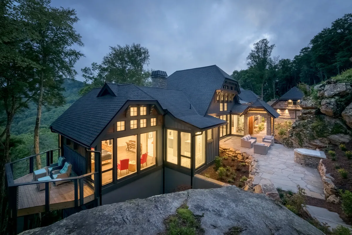 The image showcases the striking exterior of Bouldercrest, highlighting its custom architectural design with expansive windows, an inviting outdoor seating area, and a backdrop of sweeping mountain views. The scene exudes tranquility, with comfortable spaces for gathering, relaxing, and soaking in the natural beauty that surrounds the home.