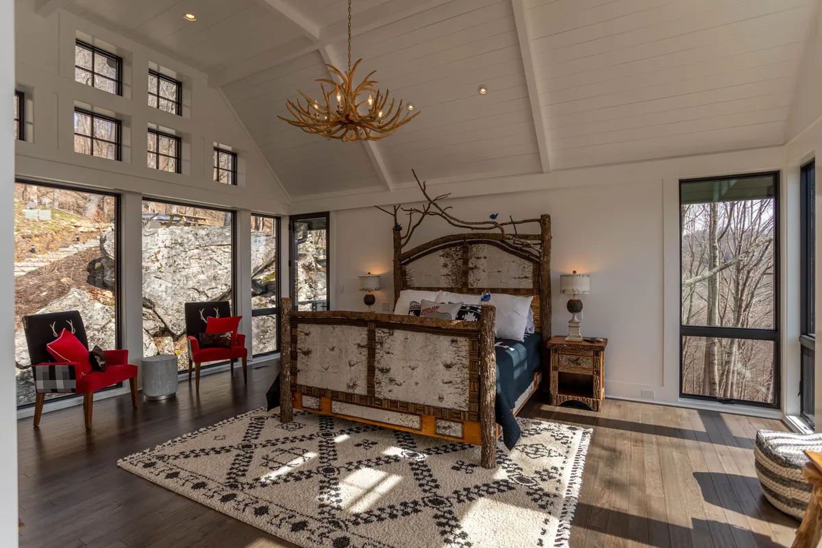 A spacious master bedroom with a wooden bed frame, chandelier, large windows offering scenic nature views, a decorative rug, and red accent chairs. The setting feels tranquil and luxurious, ideal for relaxation after a day of adventure.