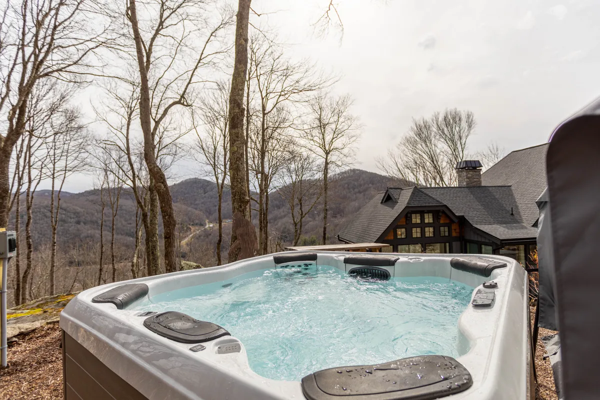 This image features an inviting hot tub positioned to overlook a breathtaking mountain view, surrounded by trees and part of the property's exterior structure. The scene suggests relaxation and socializing in nature, with plenty of space for groups to enjoy the outdoors together.