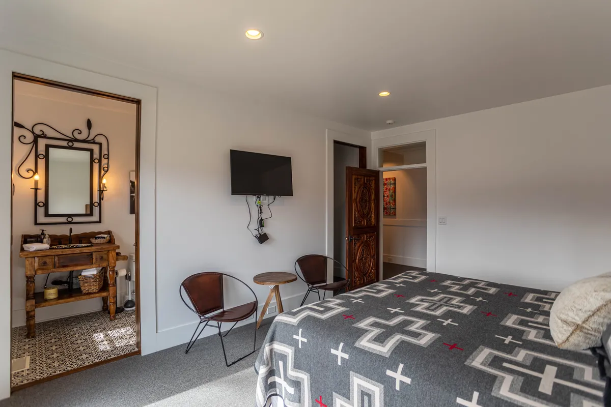 A inviting king bedroom featuring a bed with a patterned blanket, wooden vanity, decorative mirror, TV on the wall, and cozy seating in the corner. The mood is both welcoming and restful, ideal for individual guests or couples.