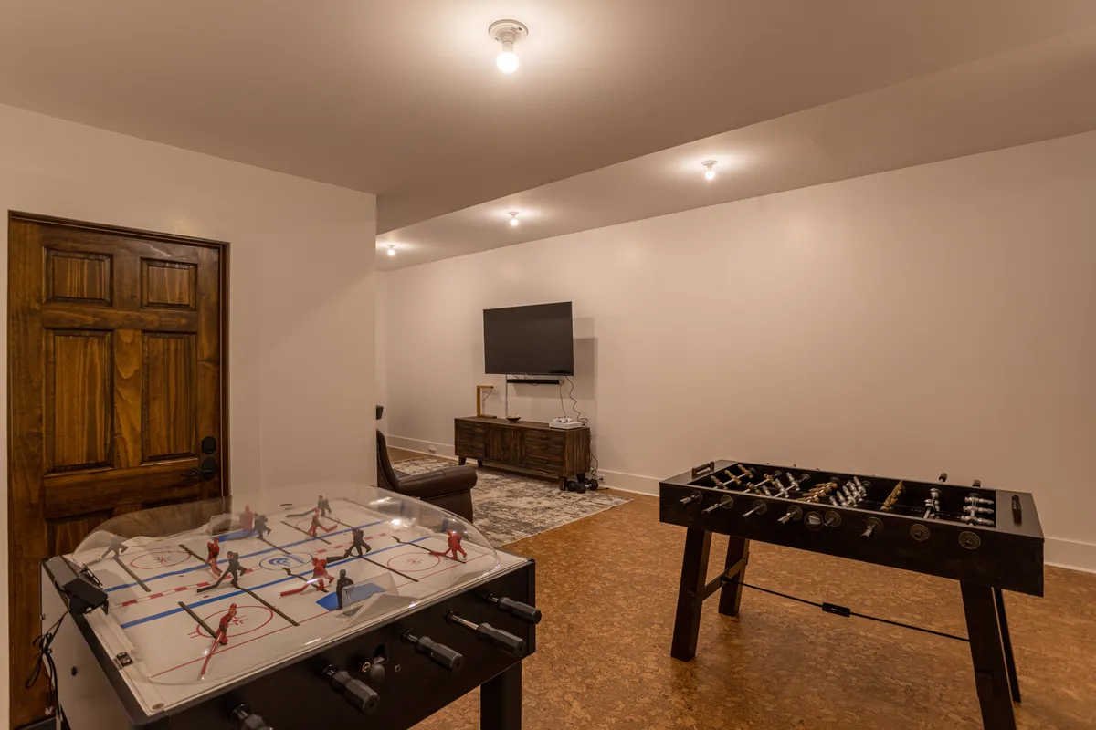 This image showcases a vibrant game room featuring both a foosball and air hockey table, a large television, and a wooden door leading to another area. The space is brightly lit and has a welcoming, playful atmosphere that invites group activities and friendly competition.