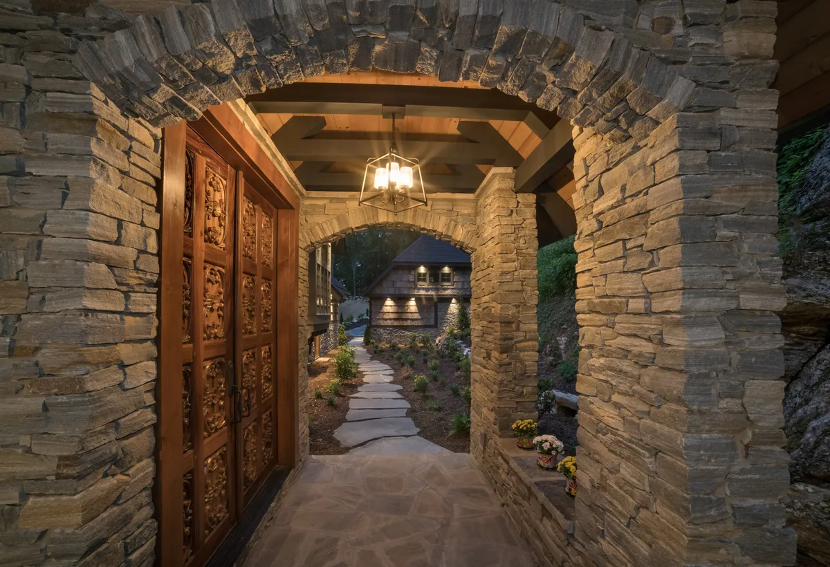 A beautifully ornate wooden door set within a stone archway opens onto a landscaped pathway, framed by lush greenery. The inviting entrance suggests both distinctive design details and a sense of arrival at a special retreat.