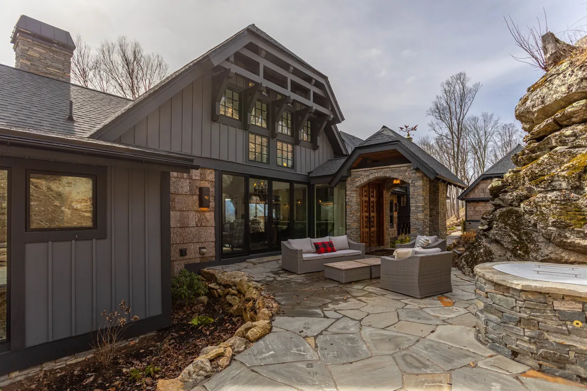 This exterior image showcases a stone patio with a comfortable seating area, a wooden entrance, and natural rock formations. The scene is surrounded by lush greenery, creating an inviting space for outdoor relaxation, gatherings, and fireside chats.