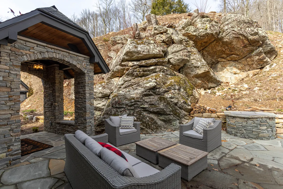 A stone wall encloses a cozy outdoor seating area centered around a fire pit. The arrangement invites guests to relax, socialize, and enjoy the warmth of the fire while surrounded by nature, making it ideal for evening gatherings.