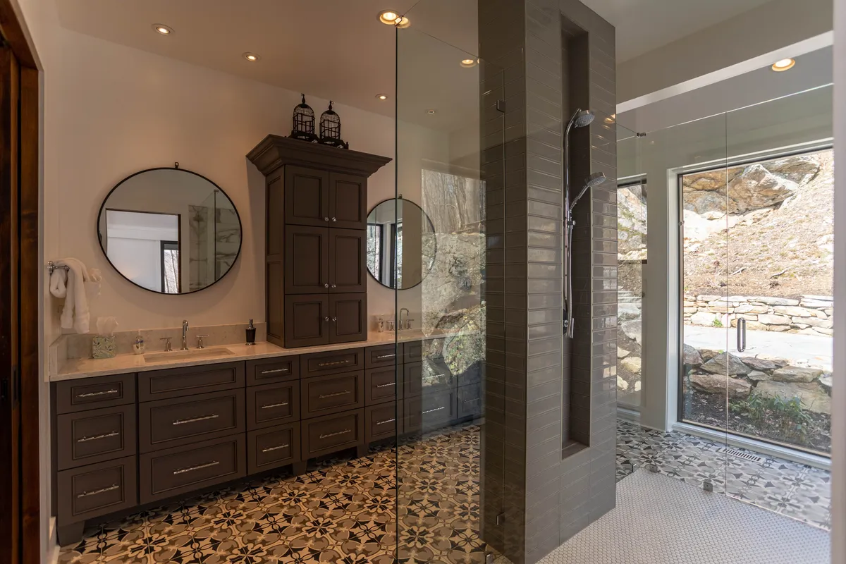 A luxurious spa-like bathroom with dual round mirrors, dark cabinetry, glass shower enclosure, and intricate floor tiles. Natural light streams in, enhancing the feeling of calm and relaxation.